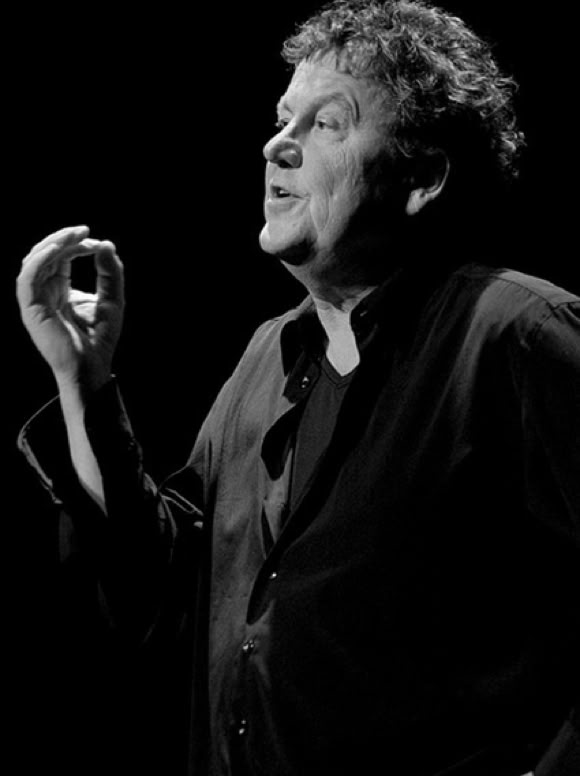 Lutz G&ouml;rner in schwarzer Kleidung gestikuliert mit der Hand, vor einem dunklen Hintergrund.