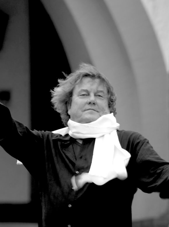 Lutz G&ouml;rner in schwarzer Kleidung mit wei&szlig;em Schal und Handschuhen, h&auml;lt einen Zylinderhut und einen Stock, im Freien vor einem Geb&auml;ude.