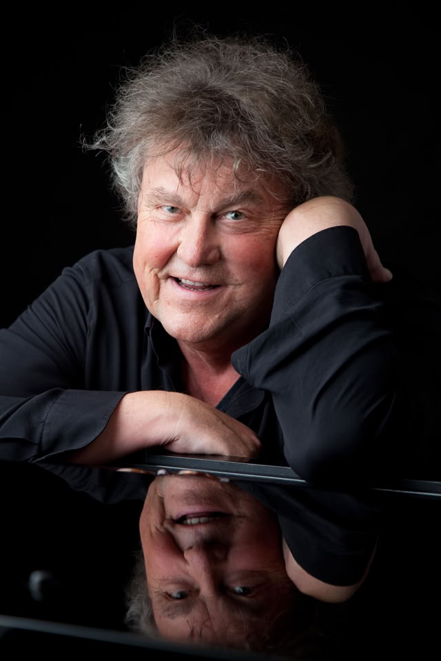 Lutz G&ouml;rner mit lockigem Haar lehnt sich auf einen Tisch und l&auml;chelt in die Kamera, mit einer Spiegelung auf der gl&auml;nzenden Oberfl&auml;che.