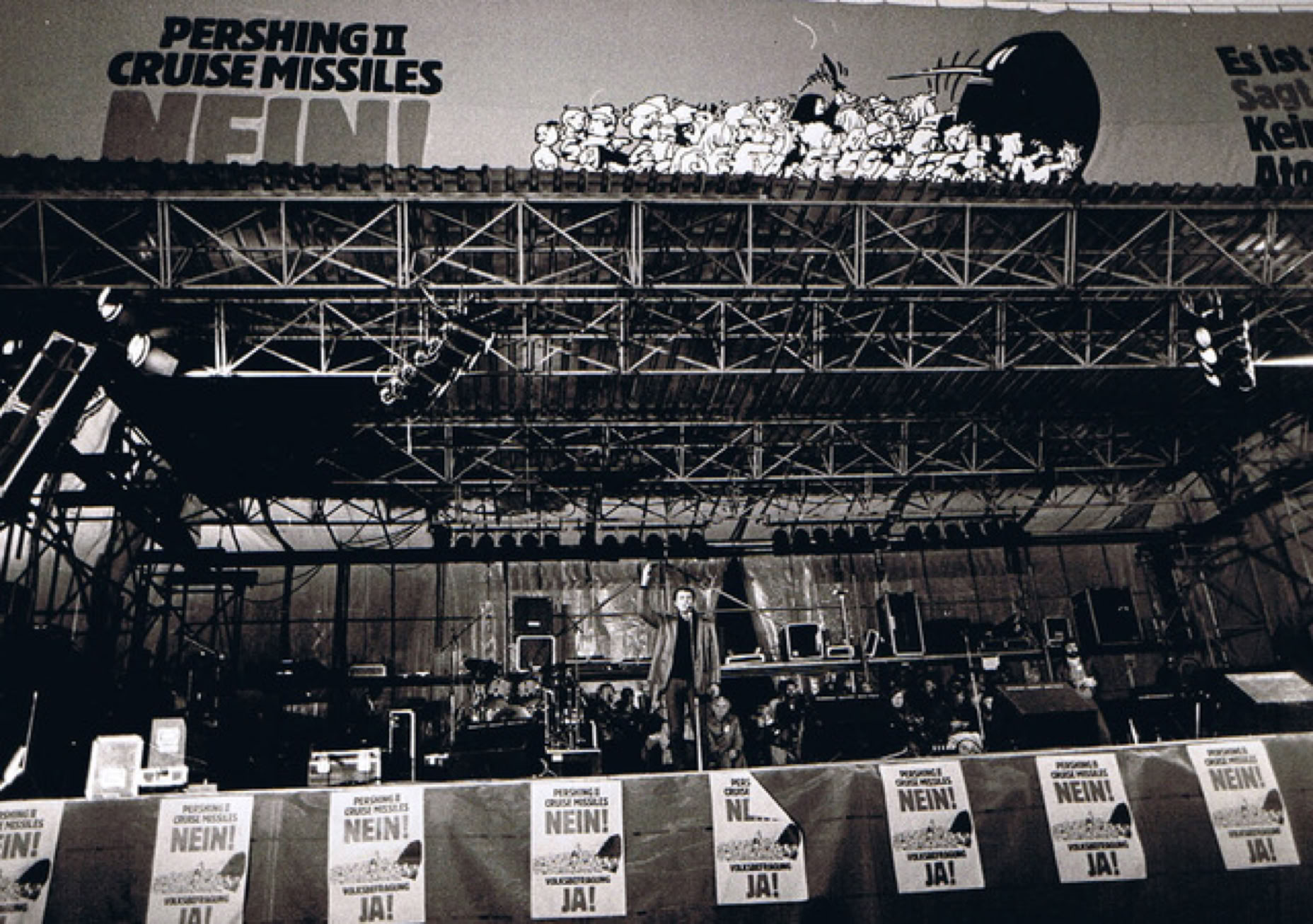 Eine B&uuml;hne mit Mikrofonen und Instrumenten, im Hintergrund ein gro&szlig;es Plakat mit der Aufschrift "PERSHING II CRUISE MISSILES NEIN!" und mehreren kleineren Plakaten im Vordergrund. Friedensdemo im Bonner Hofgarten.